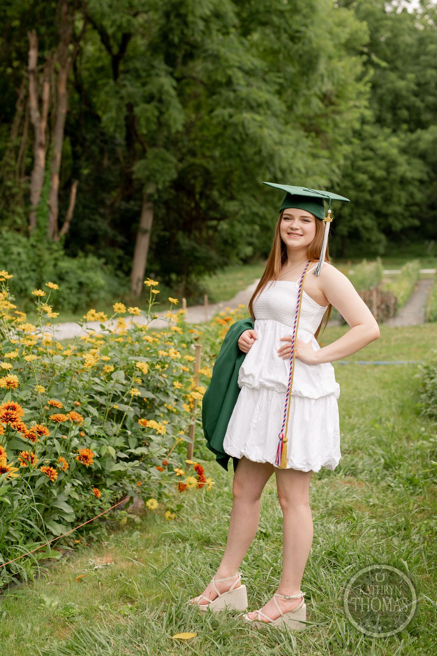 Graduate in cap and gown stands along garden path with blooming orange flowers.