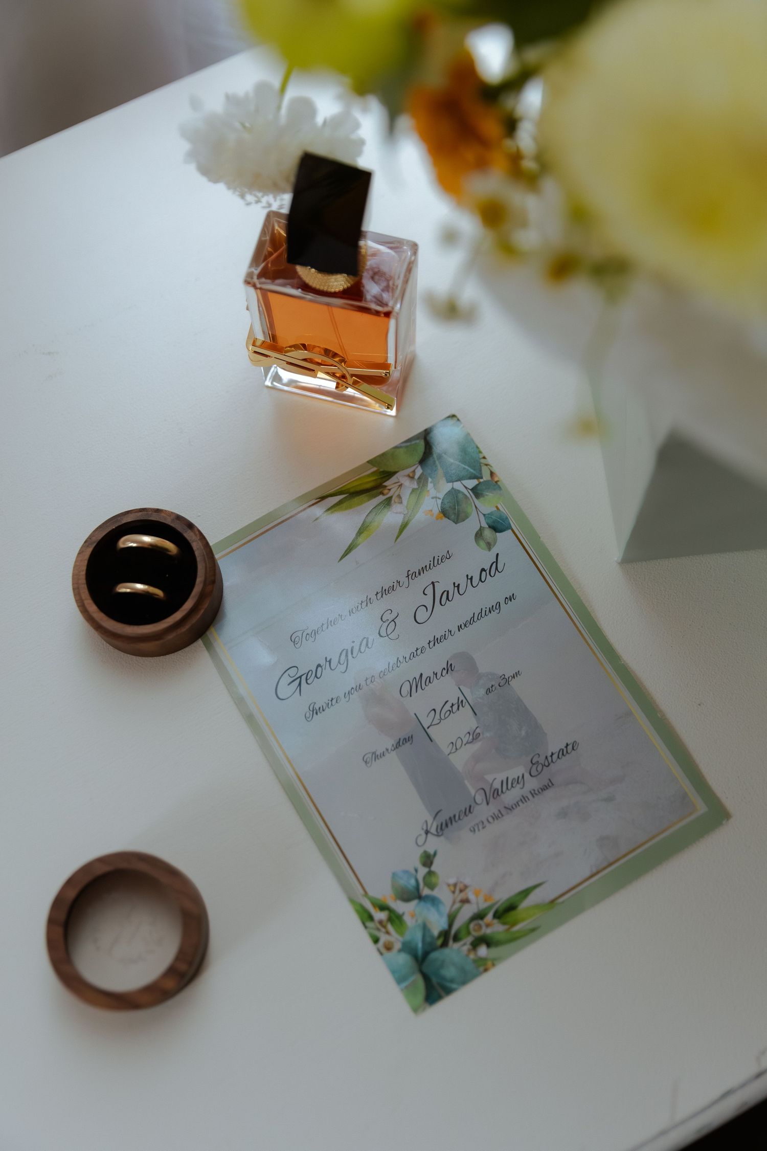 Wedding invitation for Georgia and Jarrod displayed with gold rings in wooden box and amber perfume bottle on white table.