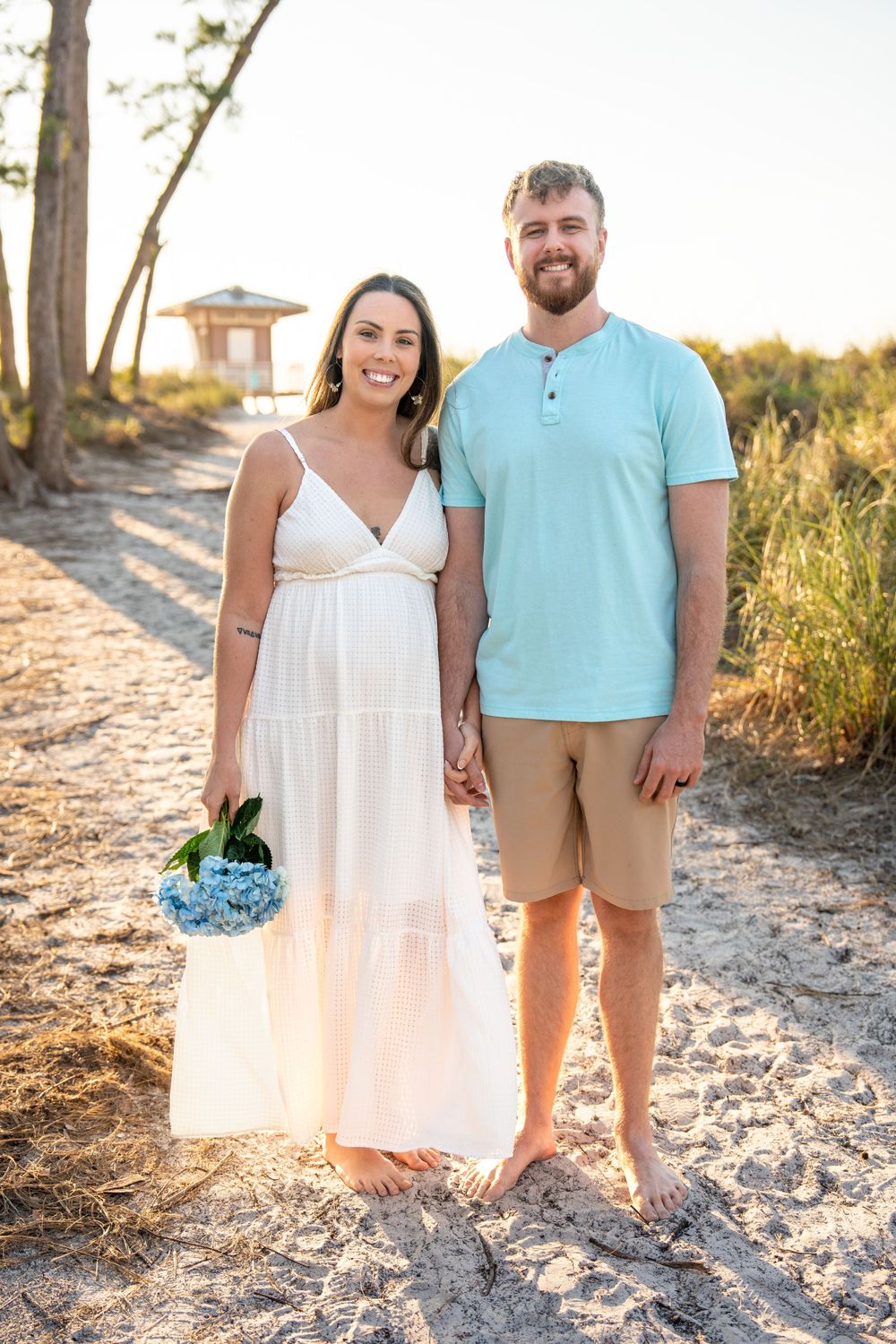 Smiling couple holding hands on sandy beach path at golden hour, woman in white dress with blue flowers.