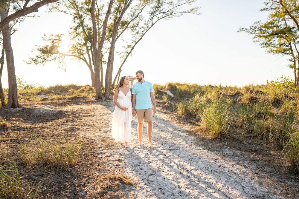 Couple walking on sandy path surrounded by trees during golden hour maternity photoshoot.