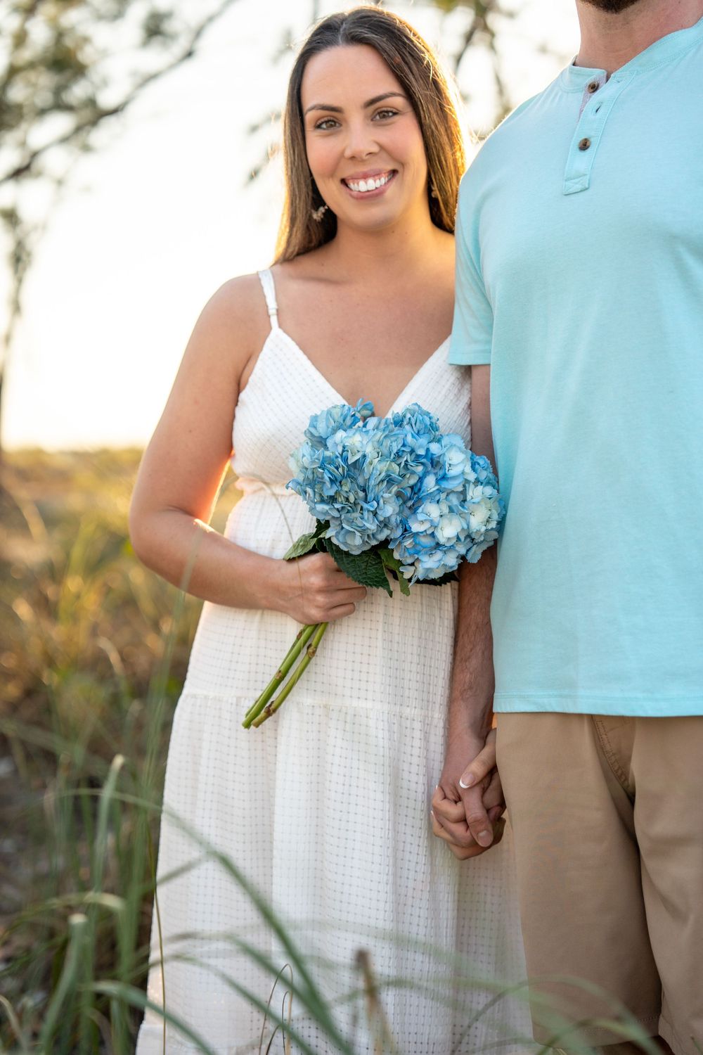 Smiling woman in white dress holding blue hydrangea bouquet at golden hour on a sandy beach path.
