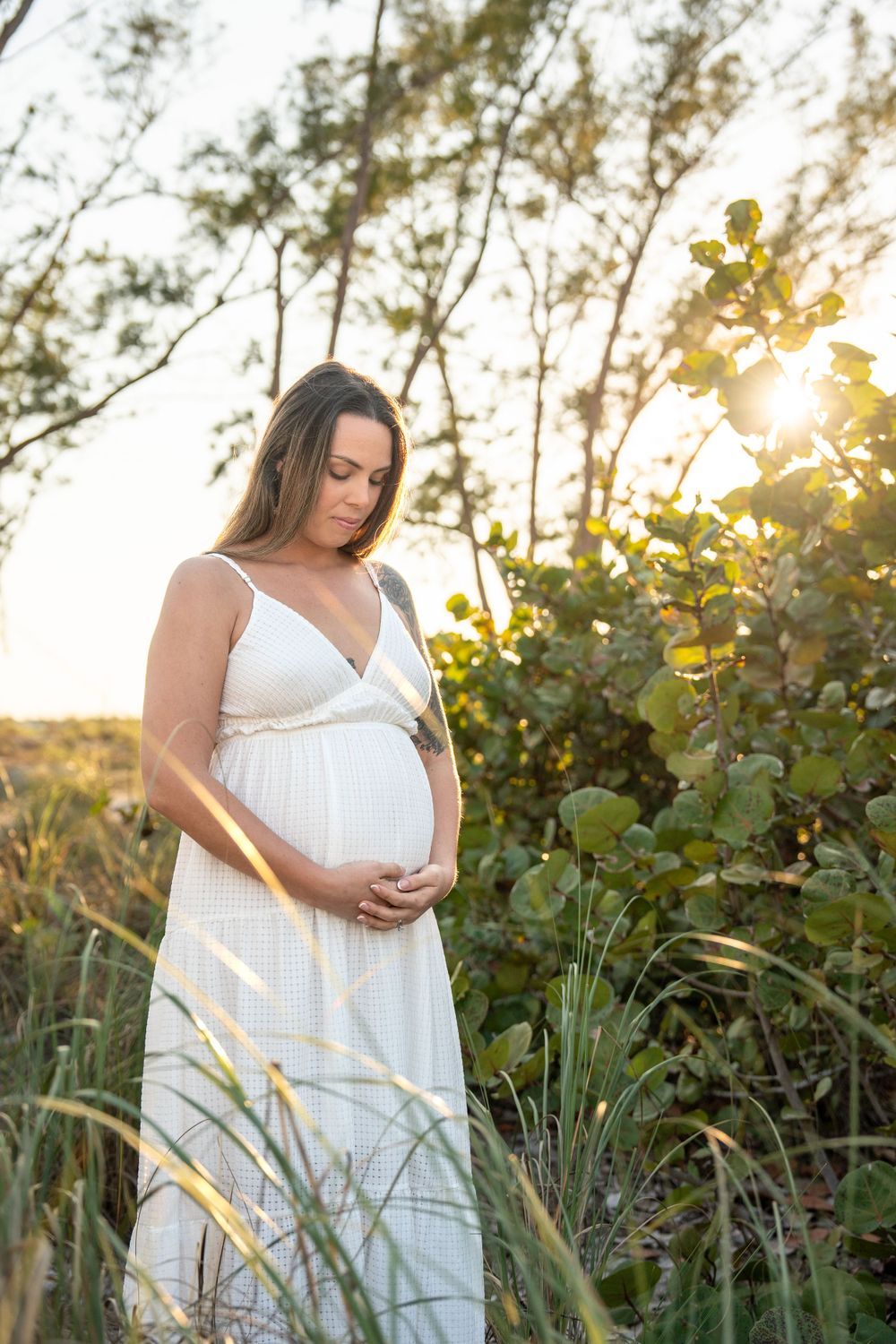 Pregnant woman in white dress cradling baby bump surrounded by lush greenery at sunset.