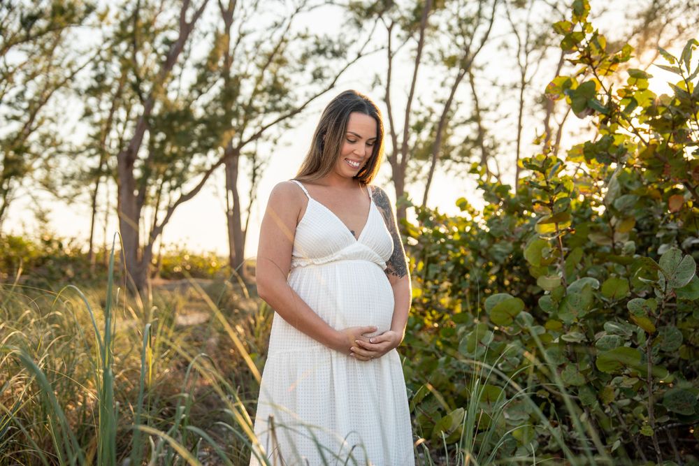 Expectant mother in white dress tenderly holding baby bump among green foliage at sunset.