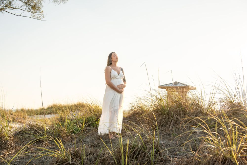 Pregnant woman in white dress standing on beach dunes at golden hour near lifeguard stand.