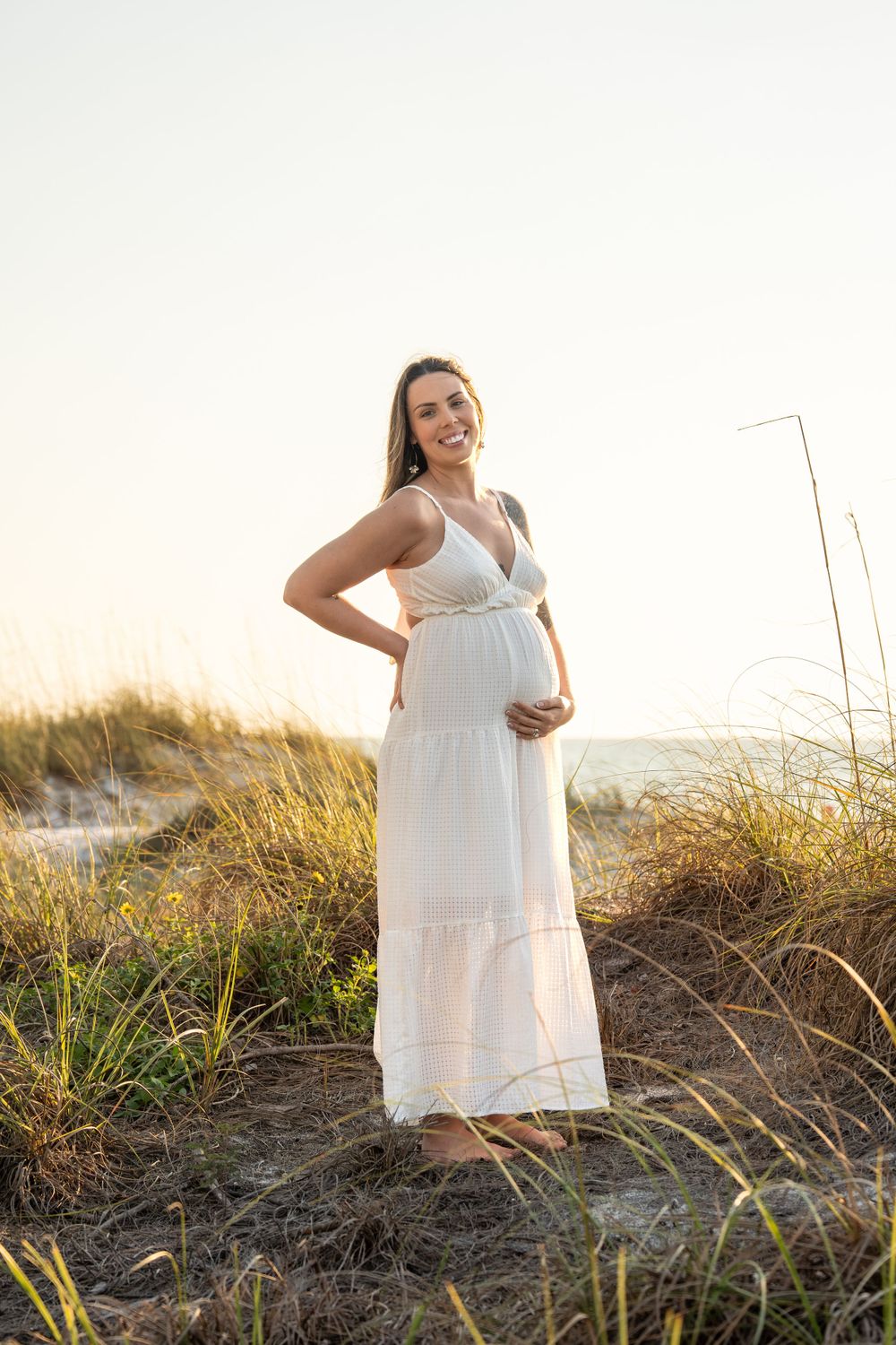 Smiling pregnant woman in white maxi dress standing in beach grass at sunset.