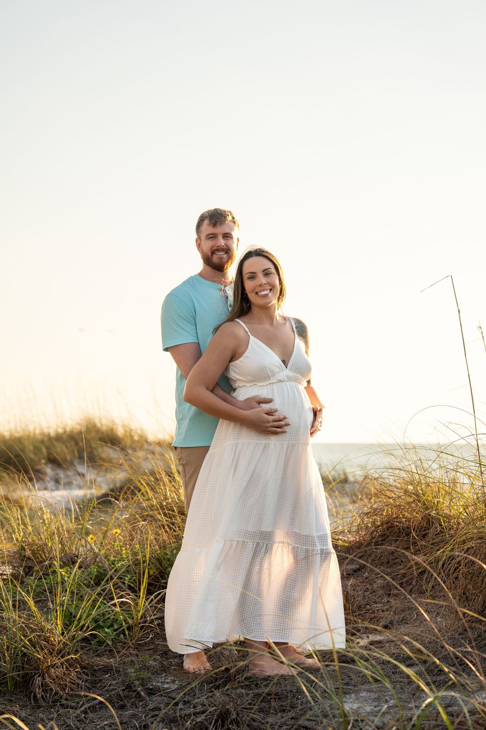 Couple embracing on beach dunes at sunset during golden hour maternity photo session.