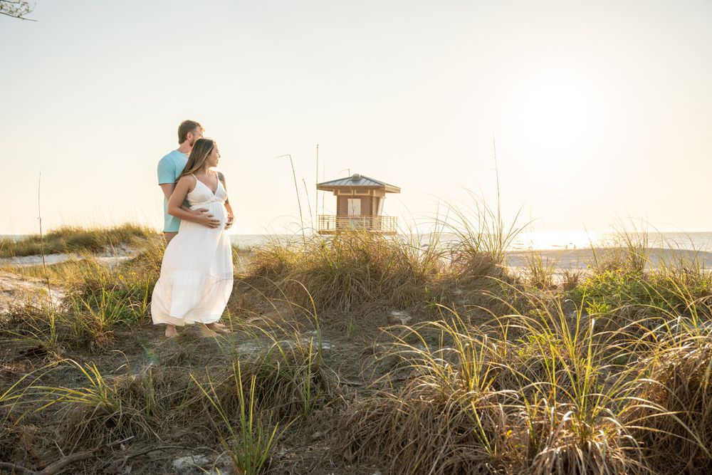 Couple embracing on sandy beach dunes near lifeguard tower during sunset maternity session.