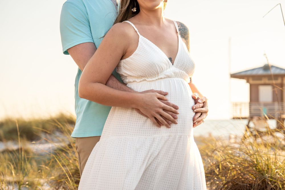 Close-up of couple holding pregnant belly together on sandy beach at golden hour.