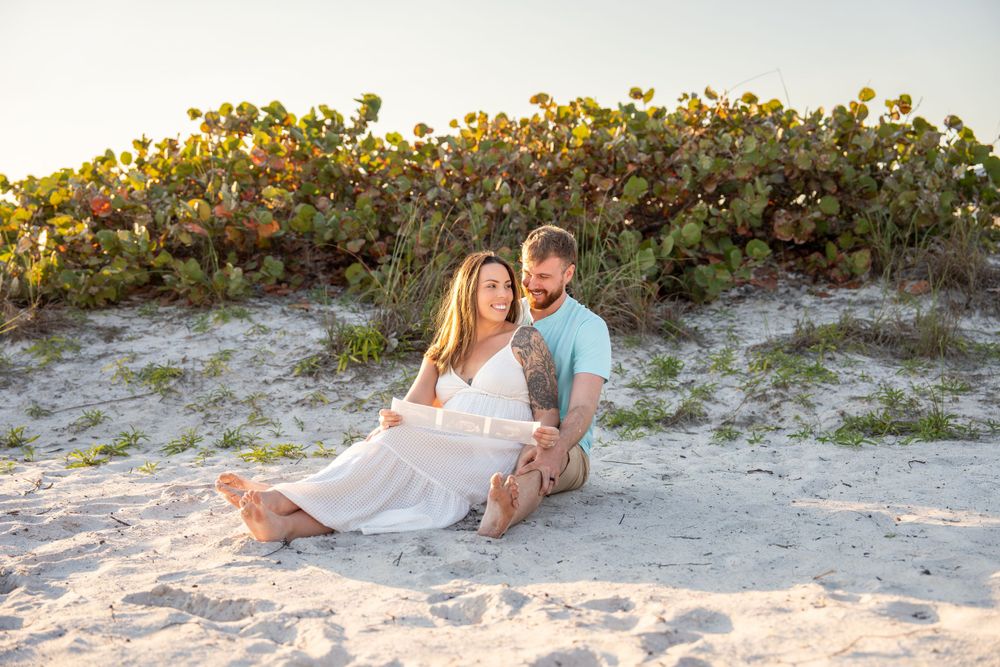 Smiling couple sitting together on white sand beach in front of colorful coastal shrubs.