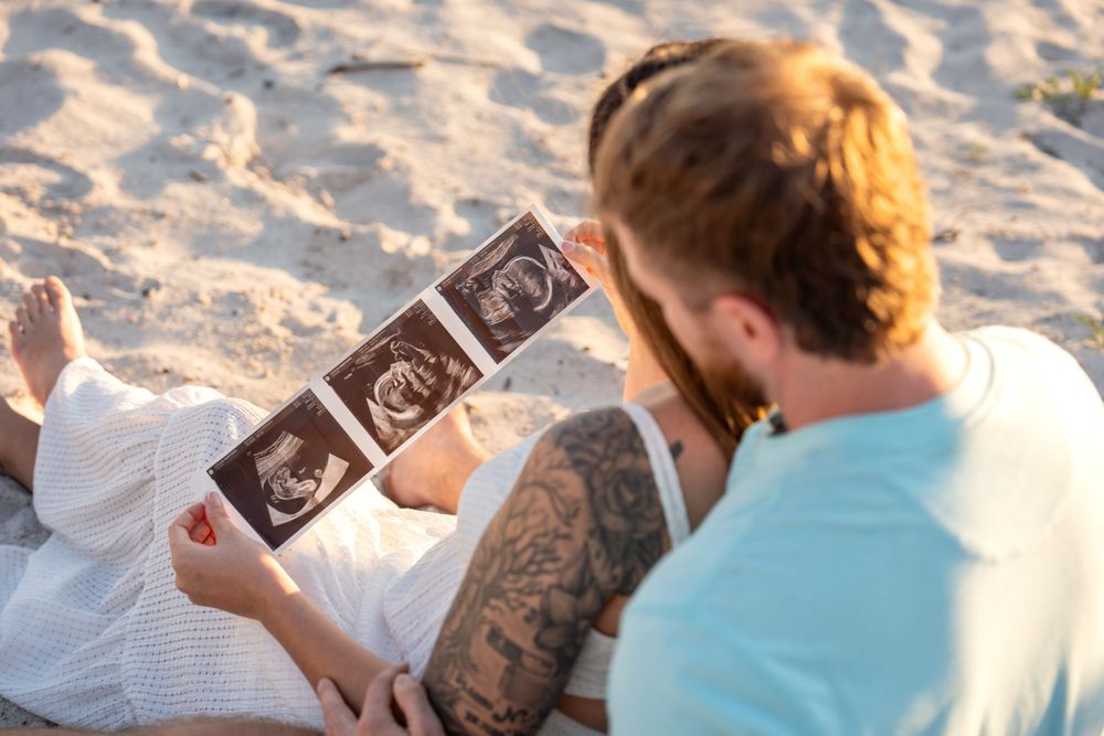 Couple sitting on sandy beach reviewing ultrasound photos during maternity photoshoot.