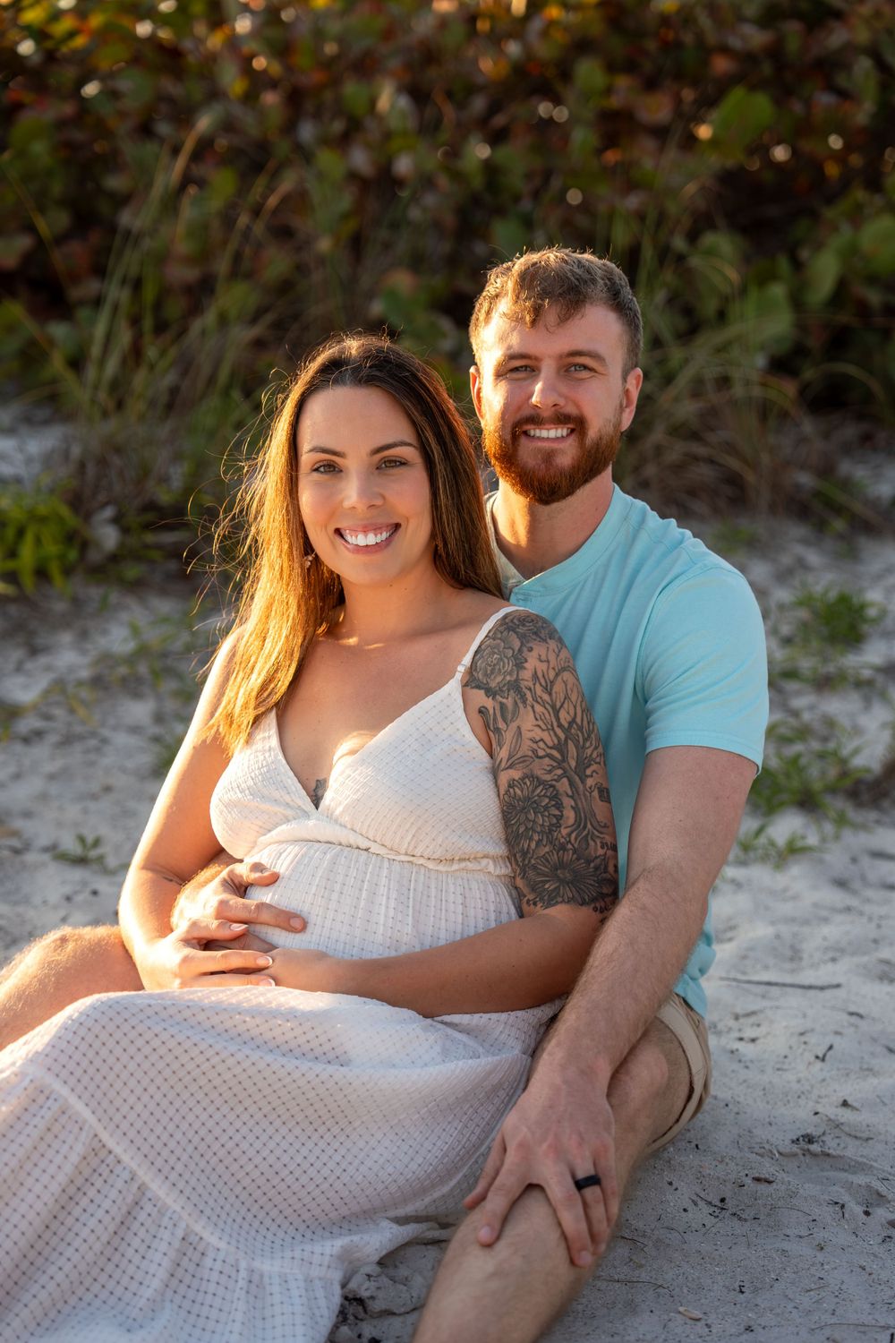 Smiling pregnant couple sitting on beach sand, woman in white dress cradling baby bump, man in teal shirt with tattoo.
