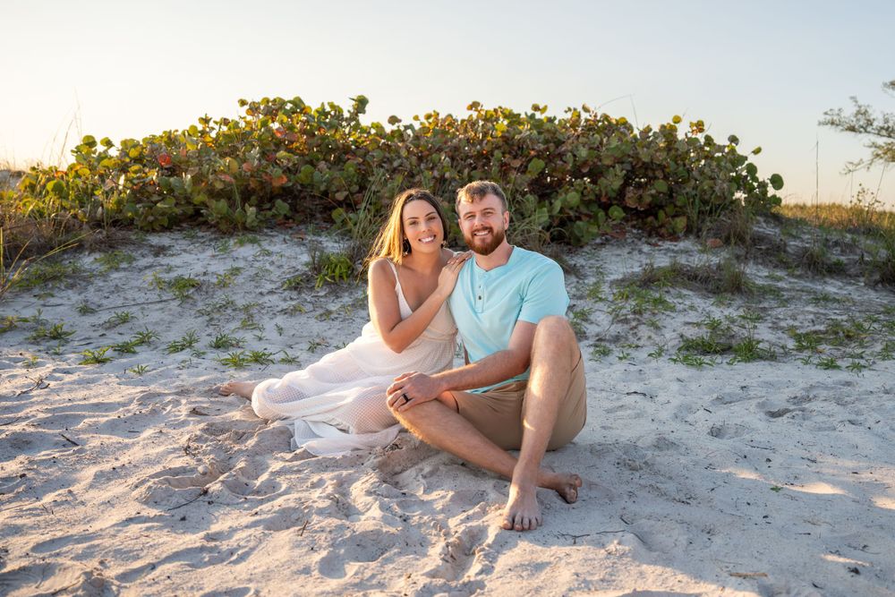 Smiling couple sitting together on white sand beach with green coastal vegetation and warm sunset light behind them.