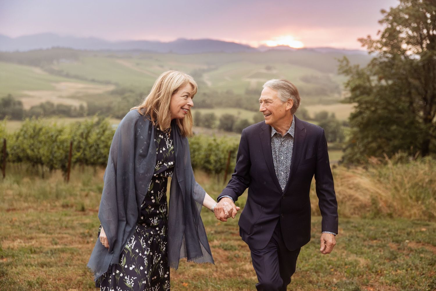 Couple walking hand in hand through vineyard at sunset against rolling hills.