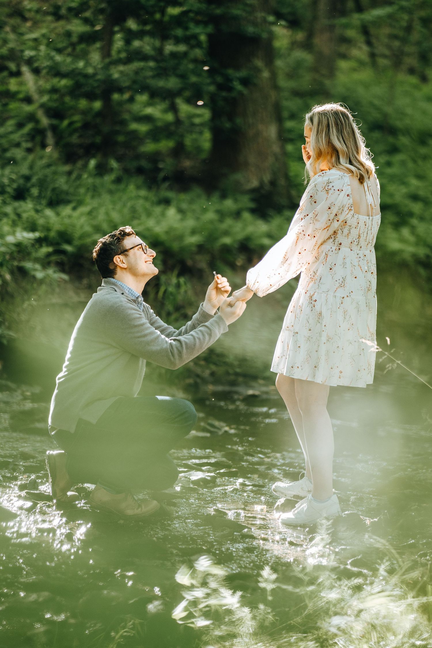 A romantic proposal moment captured in a lush green forest with soft sunlight filtering through the trees in Rockville.