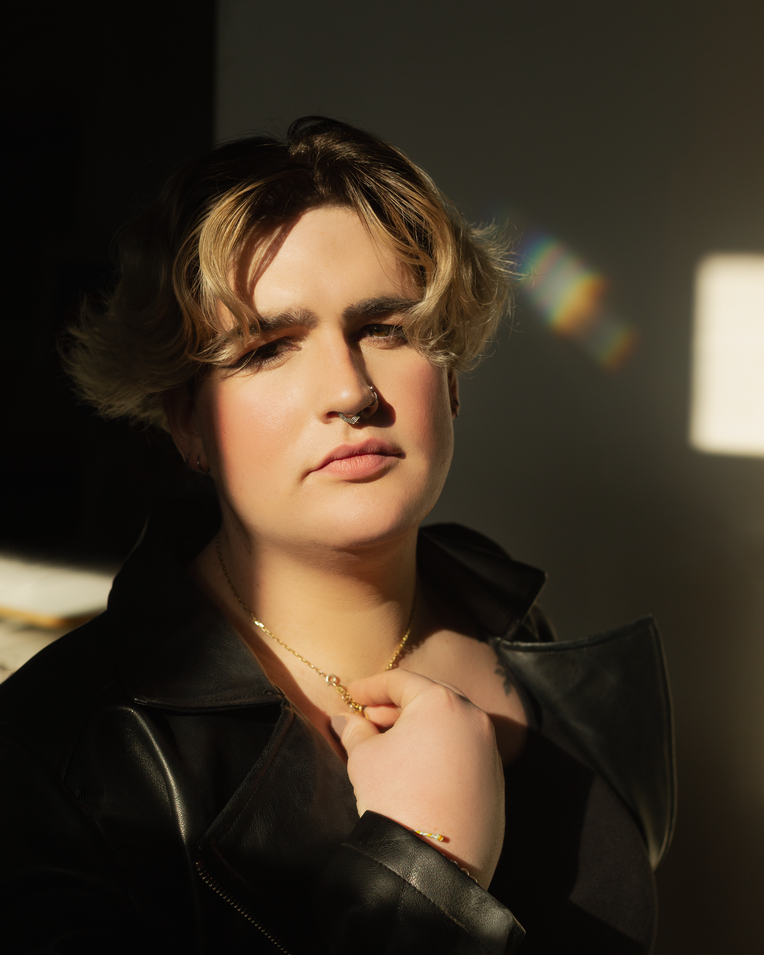 Portrait shot in warm lighting with sunbeam highlights, wearing black leather jacket, gold necklace, and wavy blonde hair.