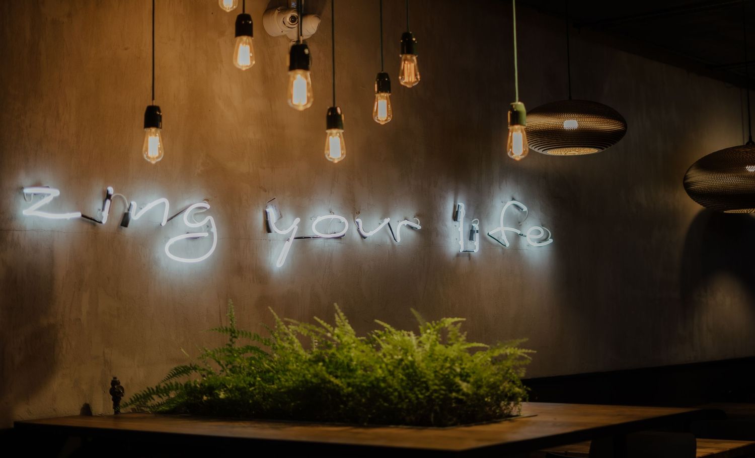 Neon sign reading 'living your life' glows above green plants with hanging Edison bulbs in moody restaurant setting.