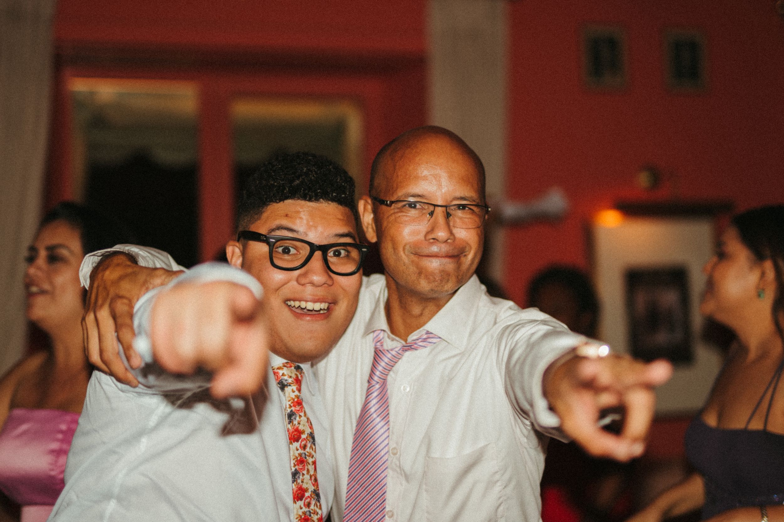 Two people at a celebration event pointing and smiling at the camera in a festive indoor setting.