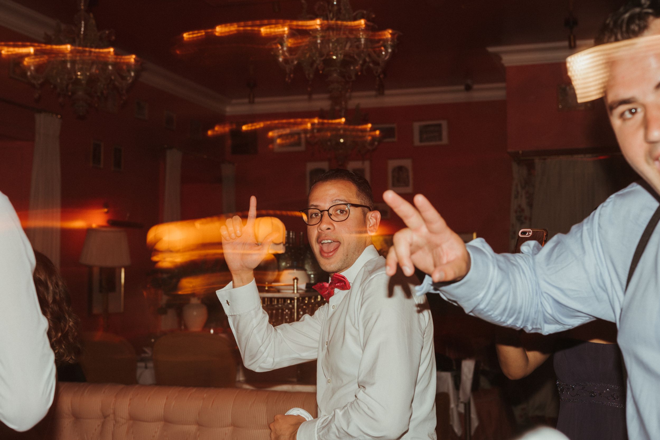 Party guests dance enthusiastically and make celebratory gestures in a red-lit venue.