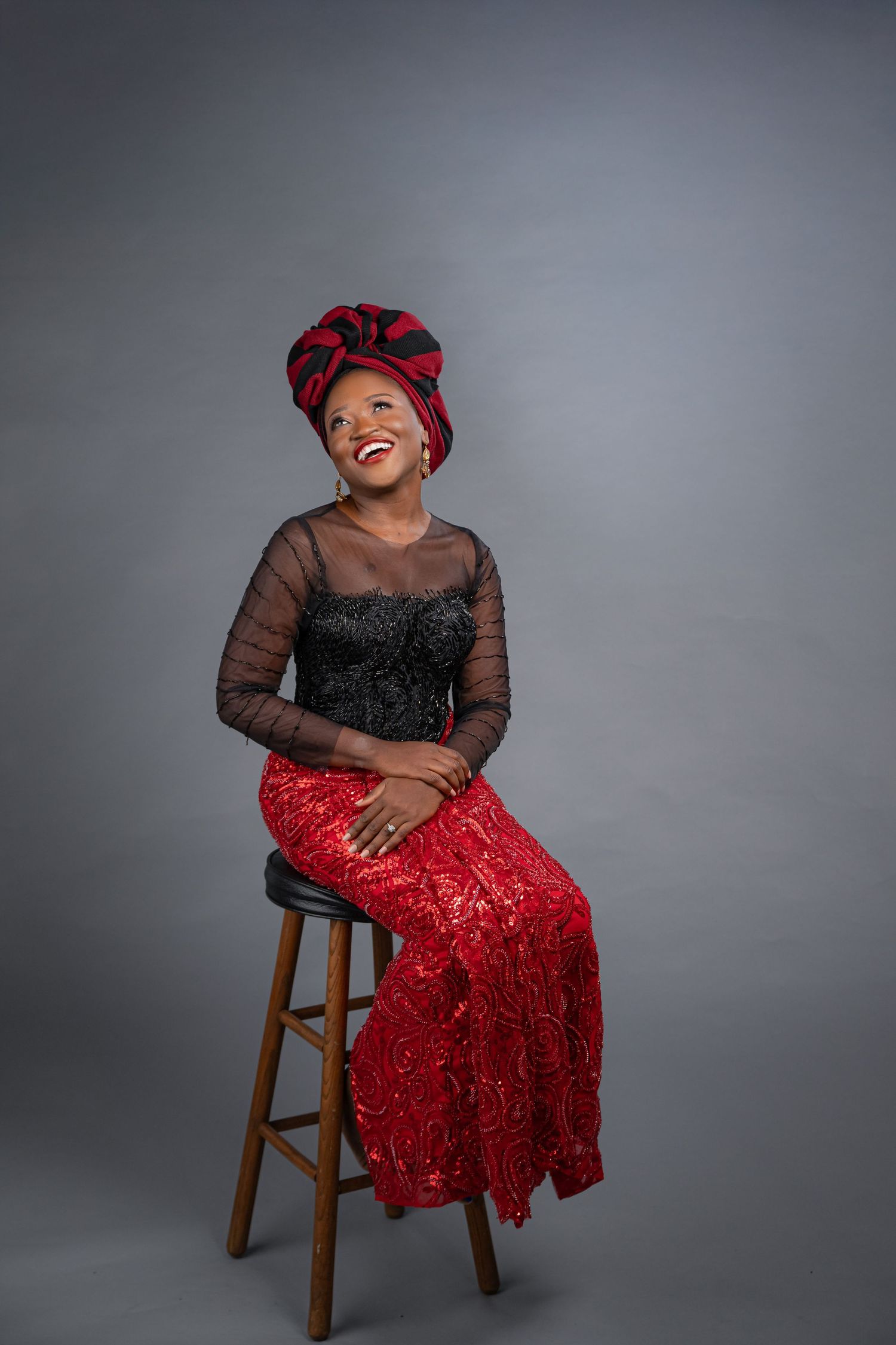 A joyful person wearing a red sequined skirt and dark sheer top sits on a wooden stool against a grey backdrop.