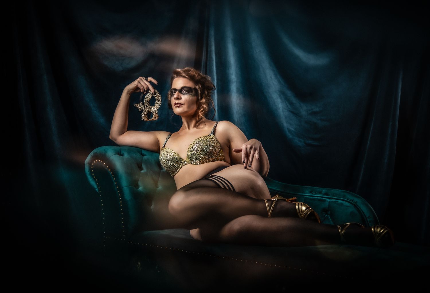Dramatic boudoir portrait on teal velvet chair with moody lighting and dark backdrop.
