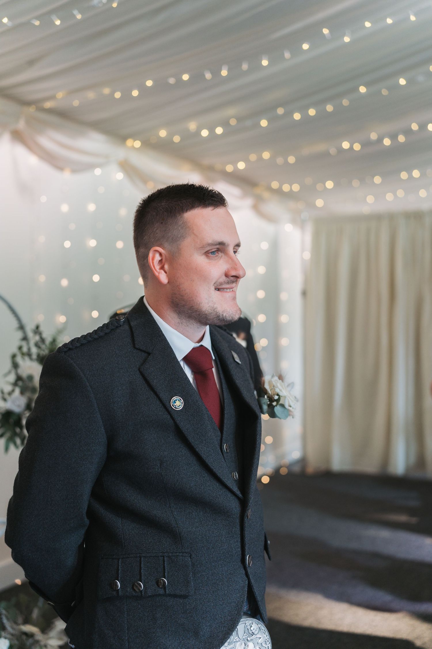 Wedding ceremony decor shows soft string lights against white drapes as someone stands in a dark suit with red tie.