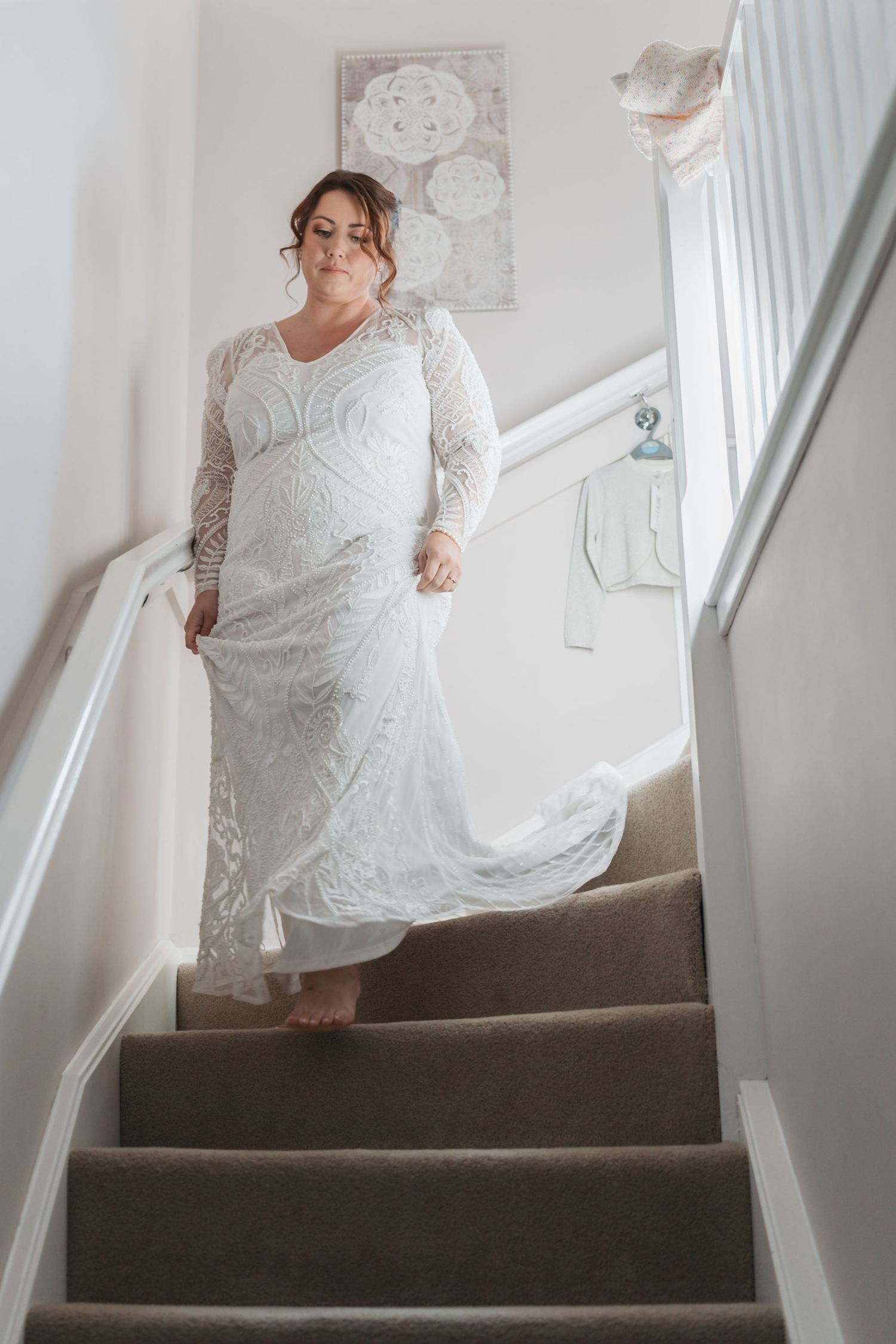 Someone wearing a beautiful long white lace wedding dress descends carpeted stairs in a bright interior hallway setting.