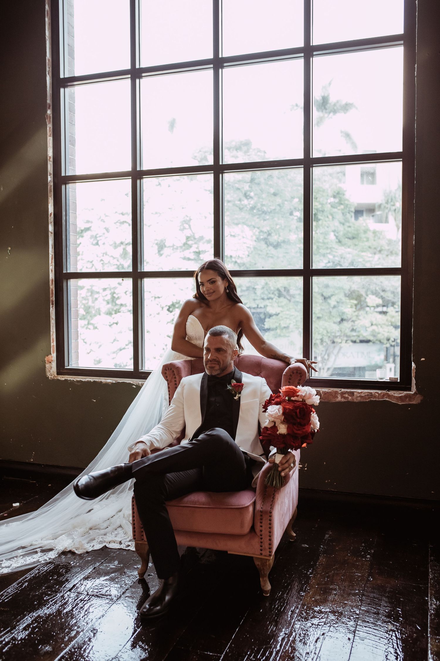 A couple poses elegantly in a rustic room with large window, seated on a vintage armchair in moody lighting.