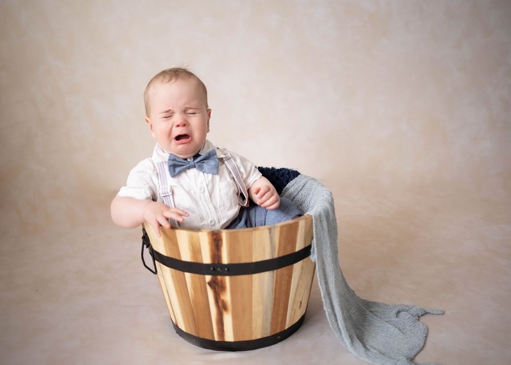 Baby sits in wooden barrel wearing blue jeans and white shirt with light blue blanket against beige backdrop.