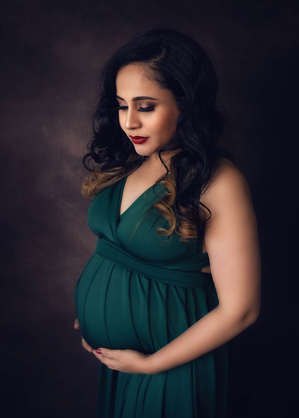 Elegant maternity portrait of woman in emerald green empire waist gown against dark background in soft lighting.