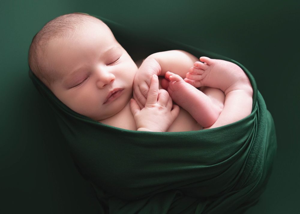 Peaceful newborn baby wrapped in dark green fabric sleeping in curled up position against green background.