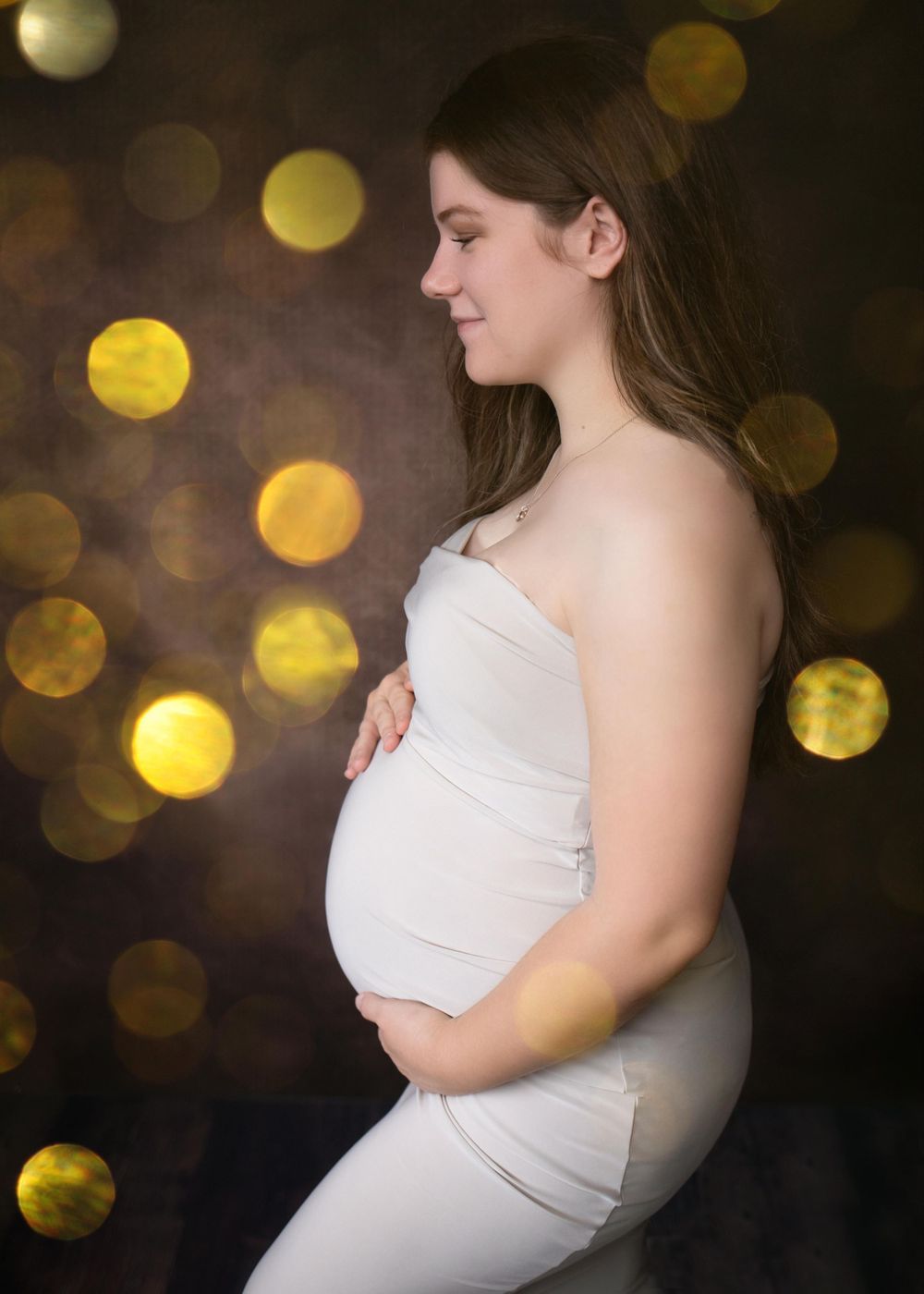 Expectant mother in white dress poses against dark background with glowing golden bokeh lights creating magical ambiance.