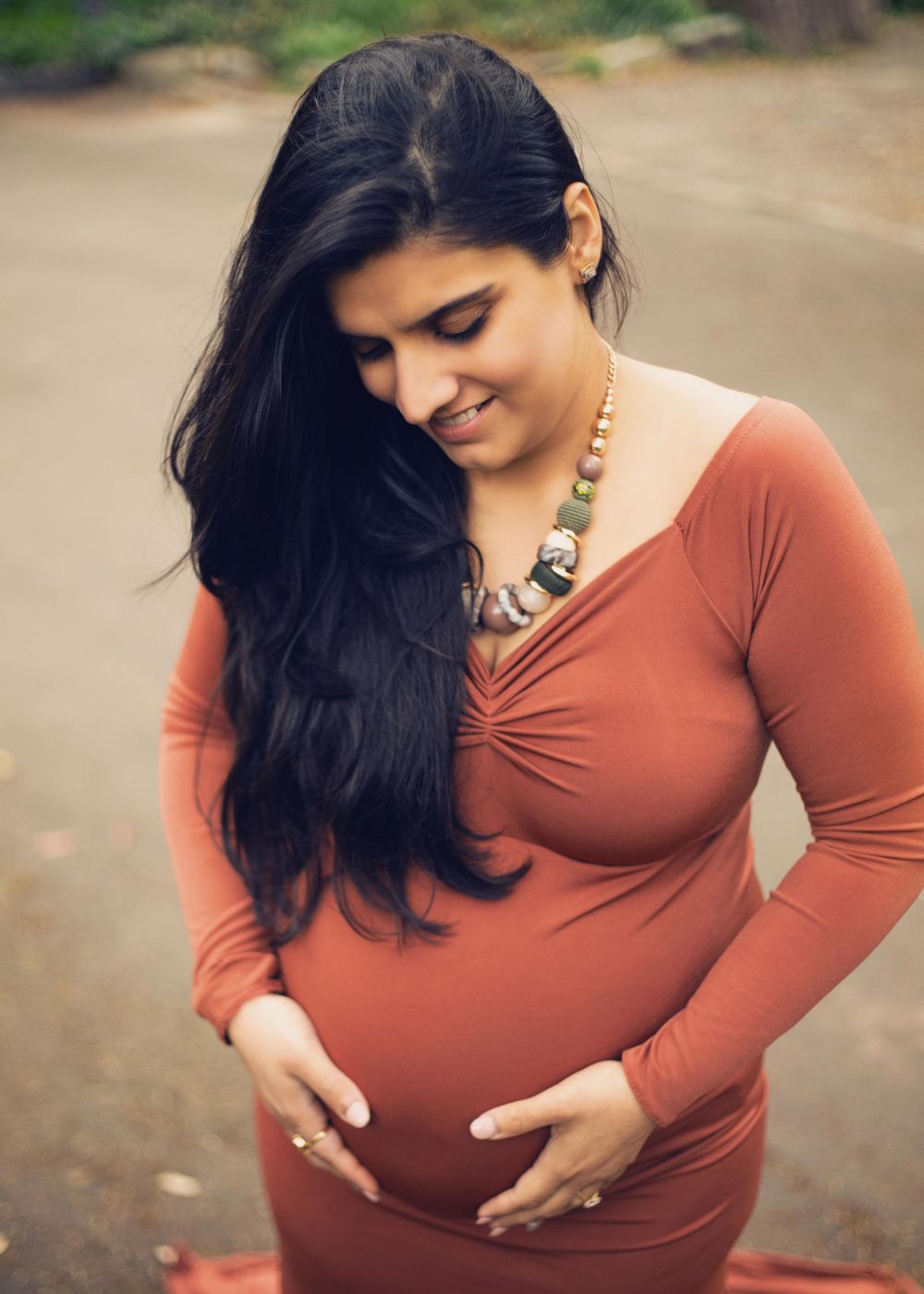 An expectant person in a rusty orange dress stands outdoors, gently cradling their pregnant belly while looking down with a smile.