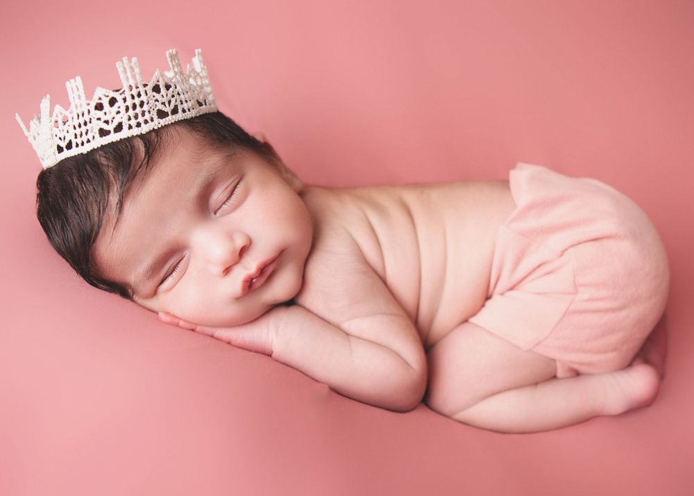 Sleeping newborn baby wearing a delicate silver crown tiara lies peacefully on soft pink background.