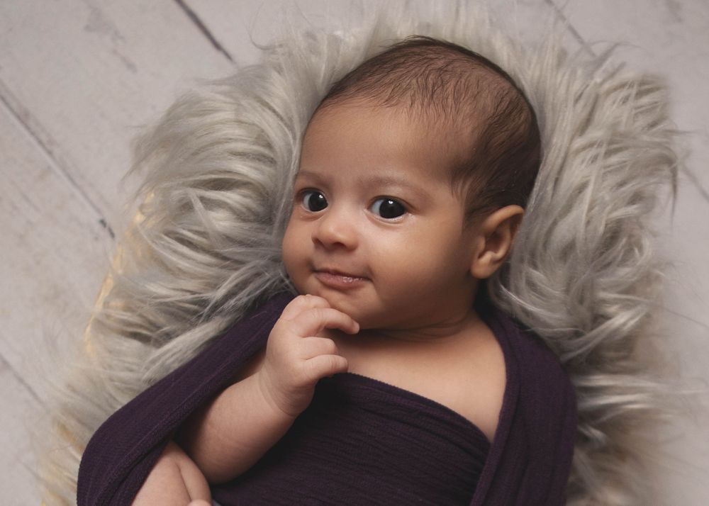 Baby with thoughtful expression lies on fluffy white blanket wearing dark purple wrap during newborn photoshoot.
