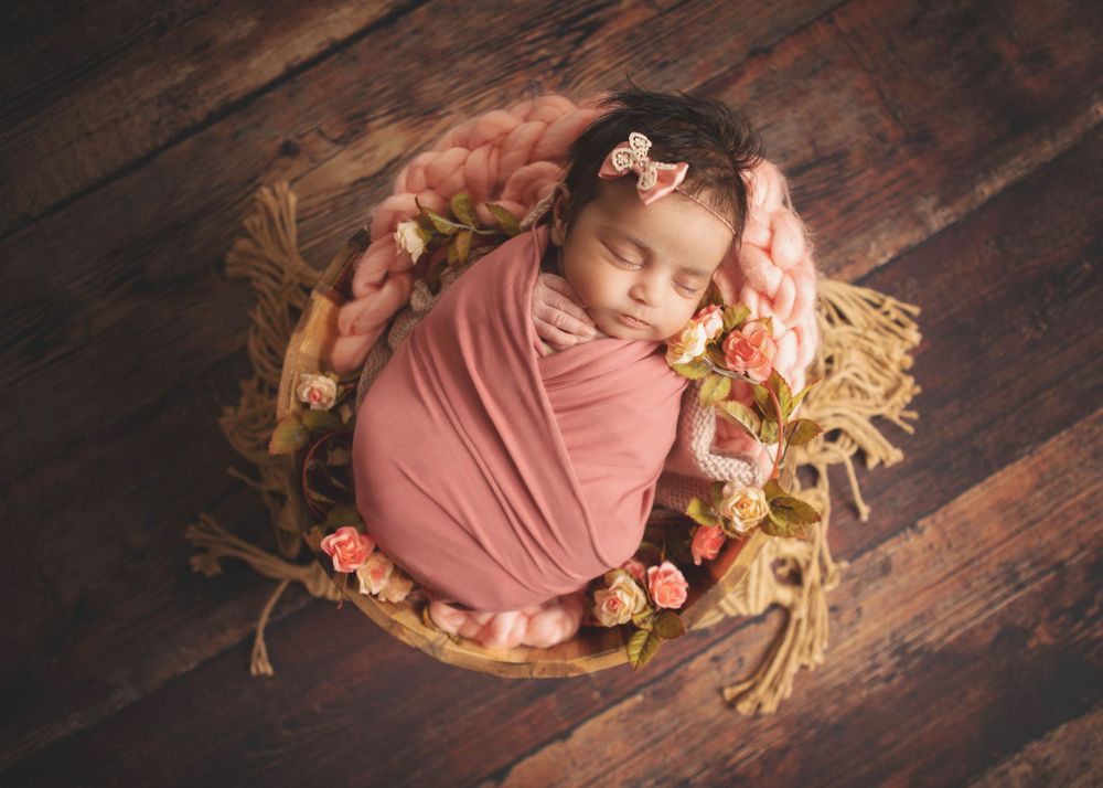 Sleeping baby wrapped in pink swaddle rests in wooden basket adorned with flowers and straw on dark wooden floor.