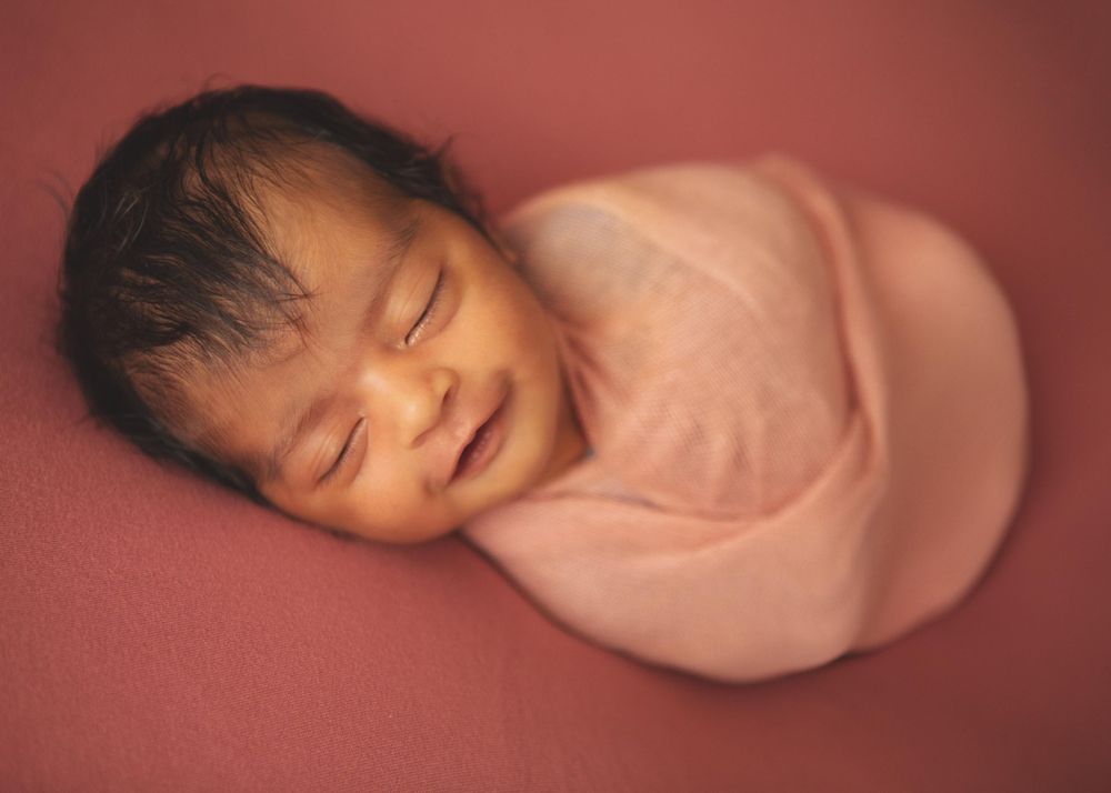 Sleeping newborn baby swaddled in soft pink fabric on pink background.