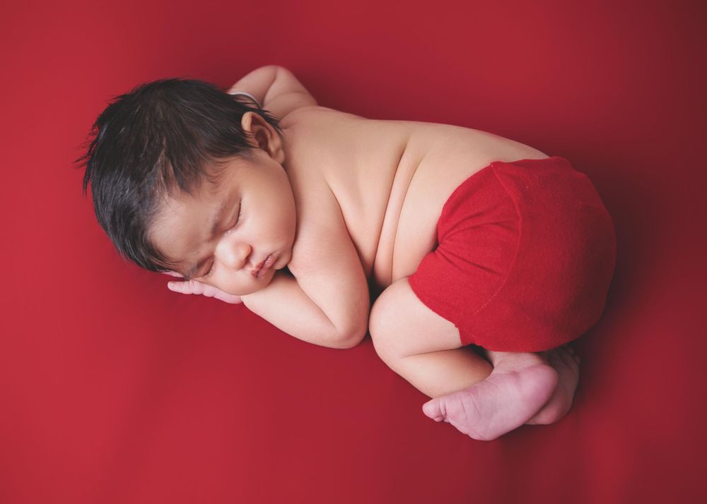 Peaceful sleeping newborn baby wearing red bottoms curled up on bright red backdrop during professional photo session.
