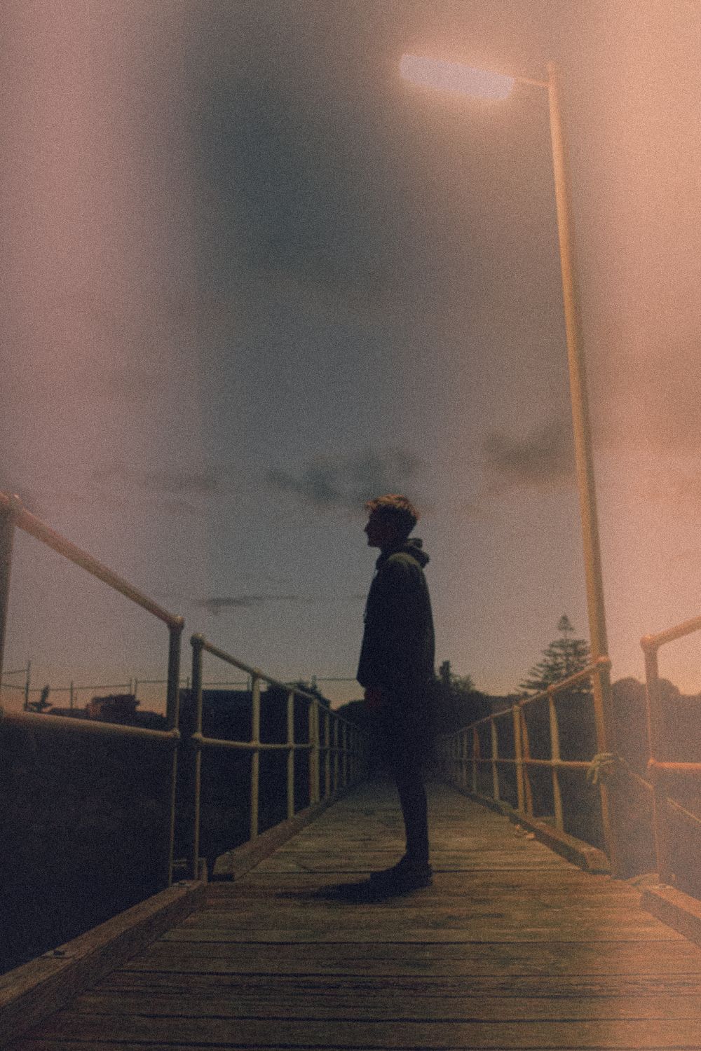 Silhouetted figure stands on a dimly lit wooden pier at dusk.