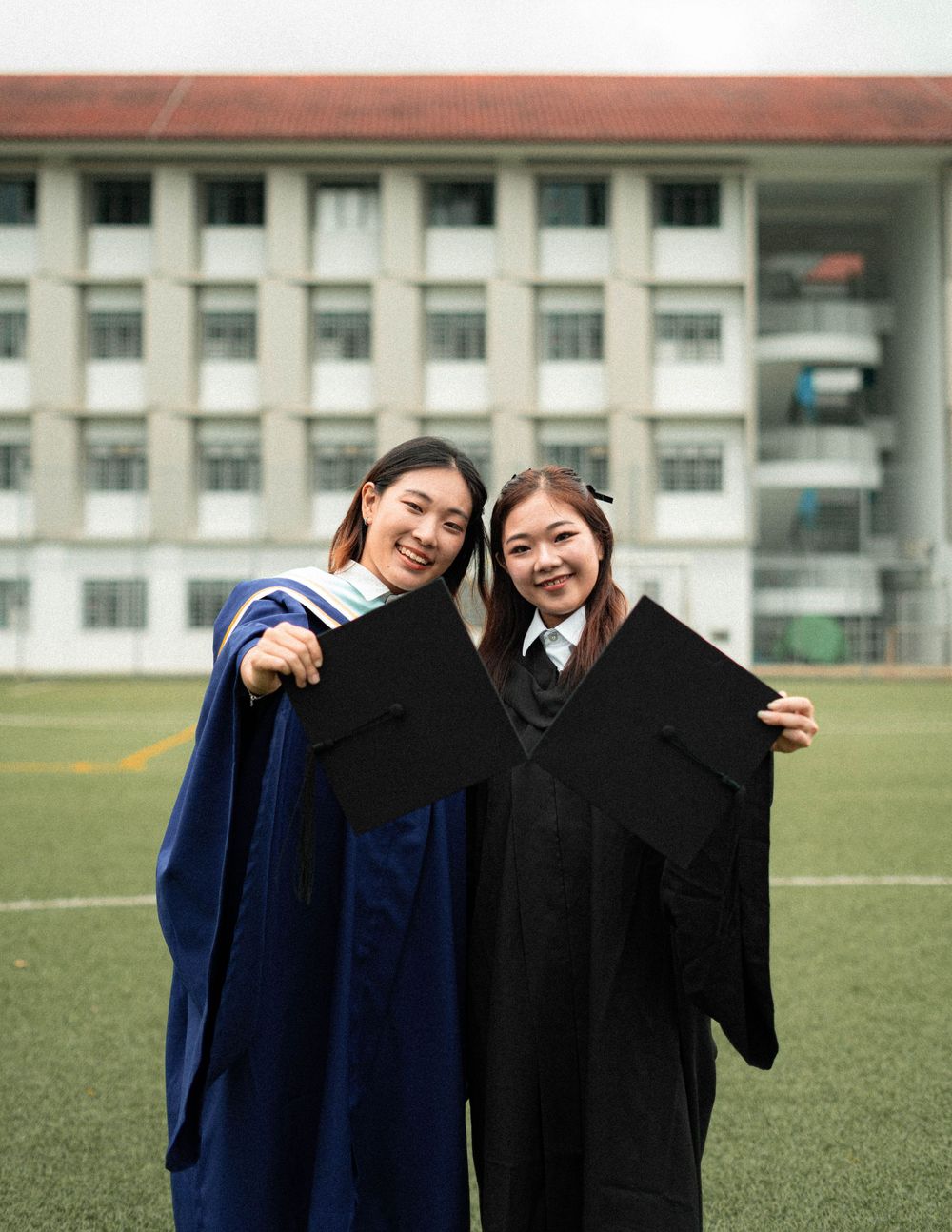 Two graduates in blue and black academic robes celebrate with their diplomas on a field.