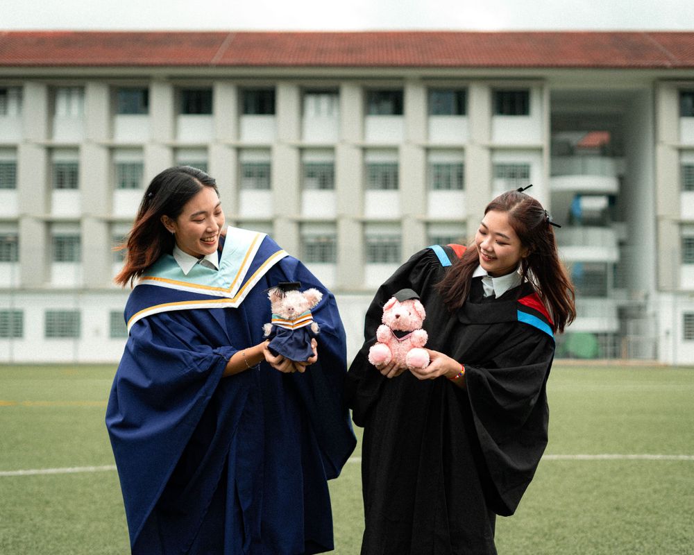 Two graduates in academic regalia hold stuffed animals while posing on a field.