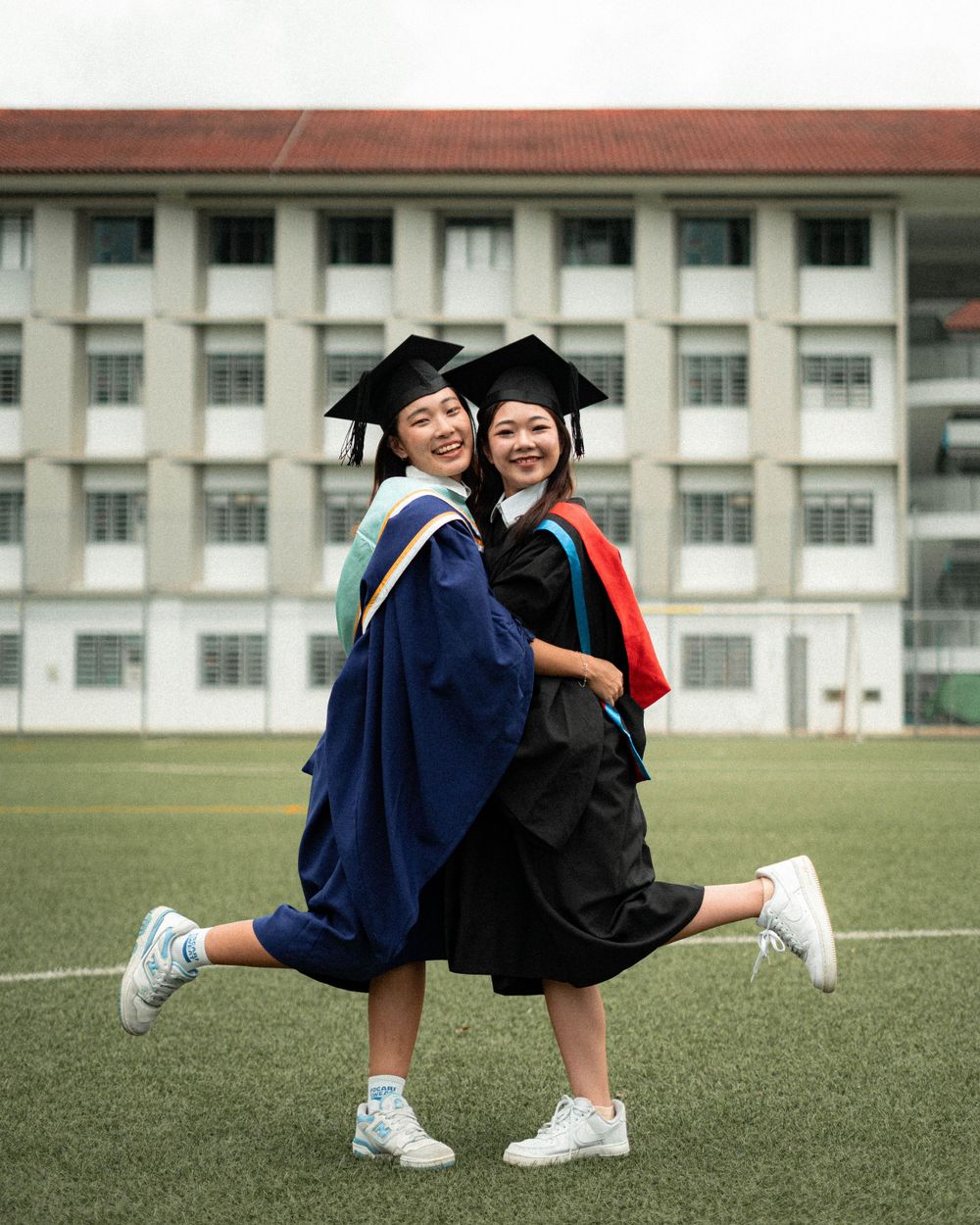Two graduates in caps and gowns strike a playful pose on campus lawn while wearing sneakers.