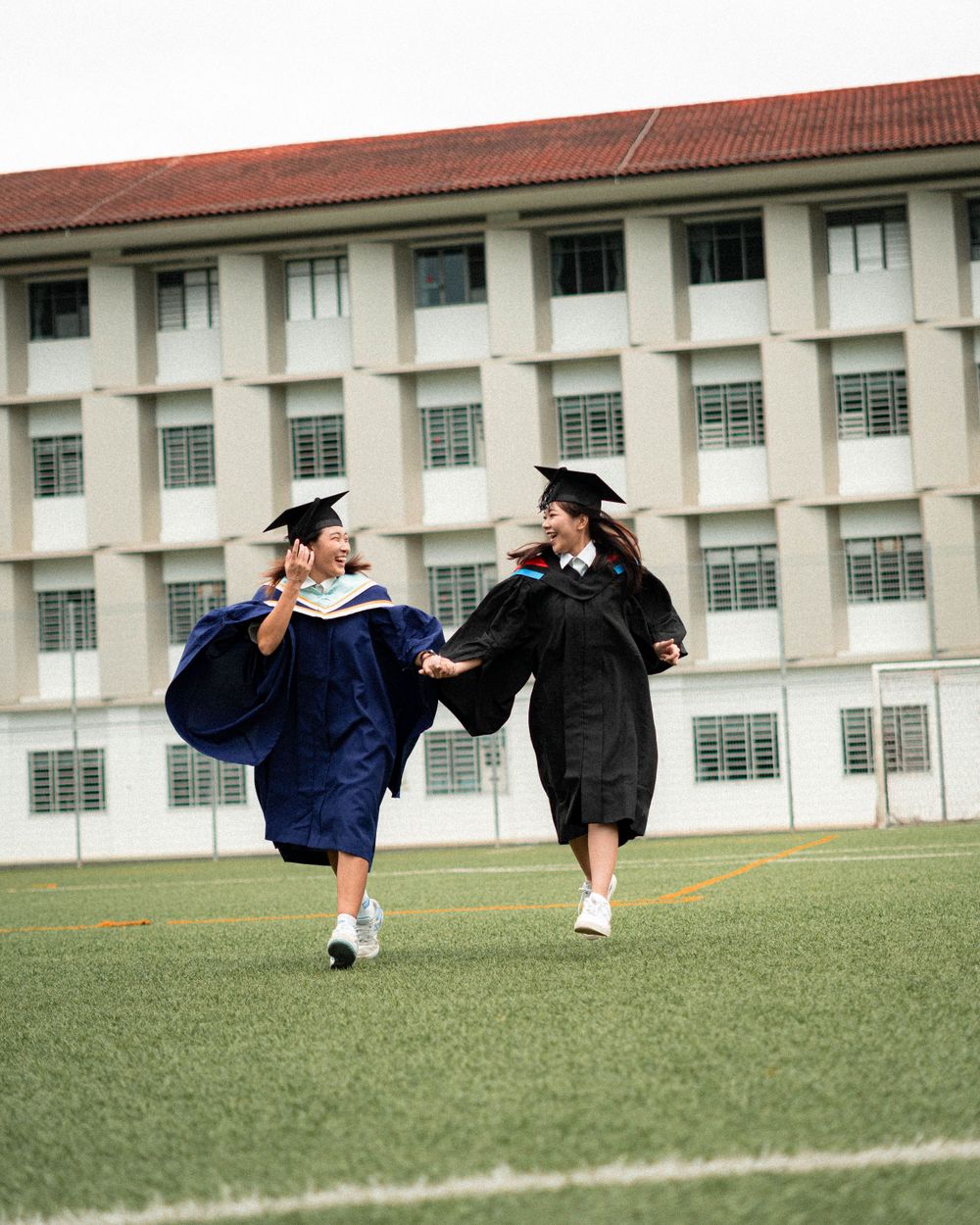 Two graduates in academic regalia dance joyfully on a grassy field with a school building in the background.