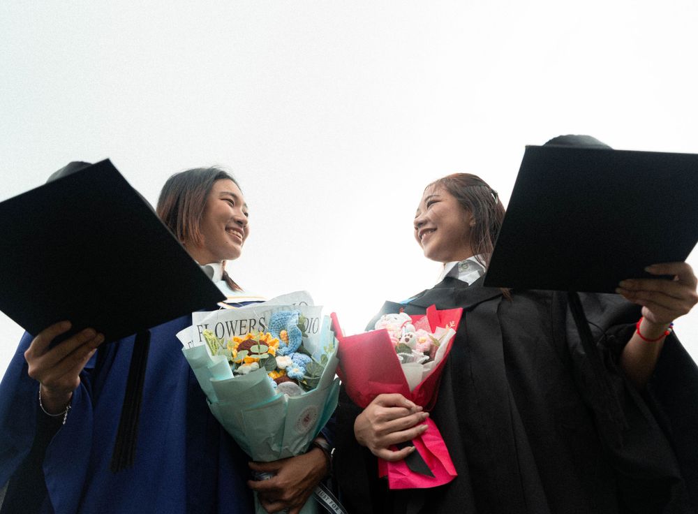Two graduates holding flower bouquets celebrate with big smiles against white sky.