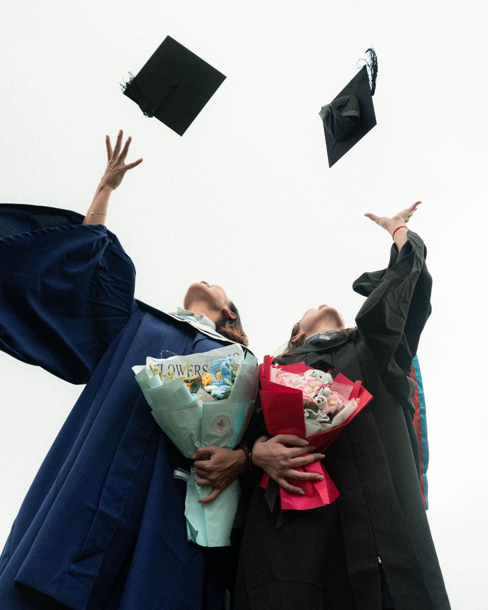 Graduates toss their caps in the air while holding colorful bouquets during graduation celebration.