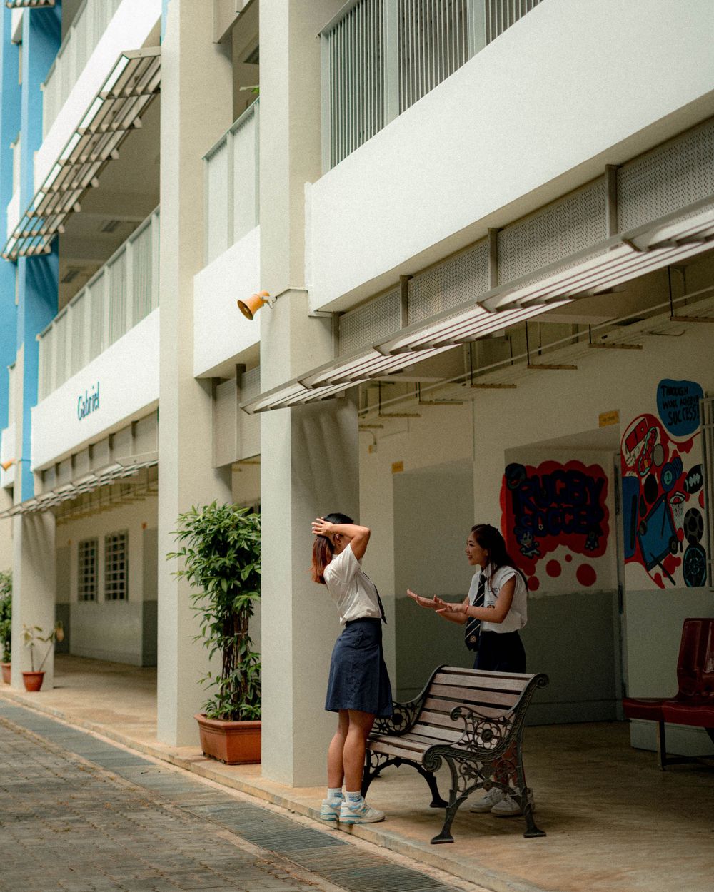 Two students in uniforms sit on a bench in front of school corridor with mural art.