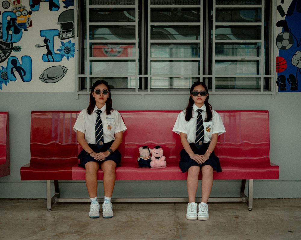 Two students in school uniforms sit on red bench wearing sunglasses.