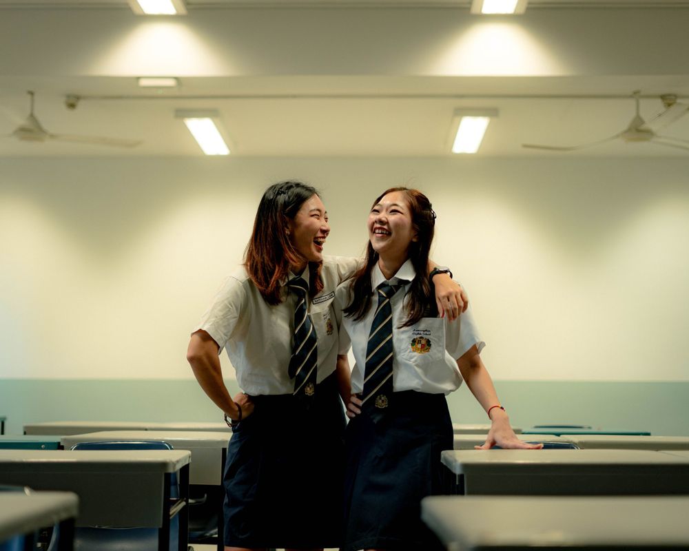 Two students in school uniforms share a lighthearted moment together in a classroom.