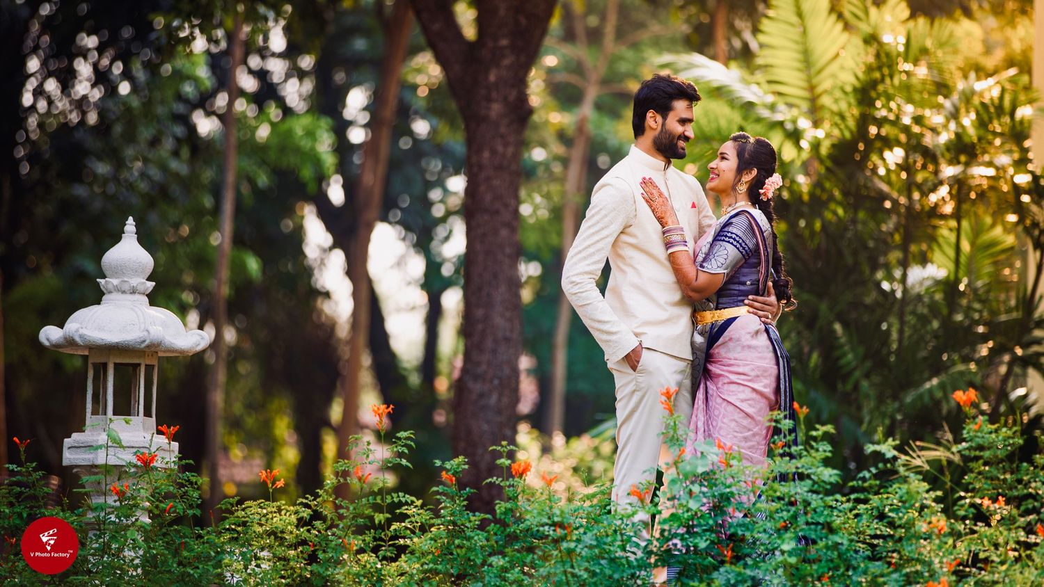 A romantic couple poses in traditional Indian attire near a garden lantern surrounded by lush greenery.
