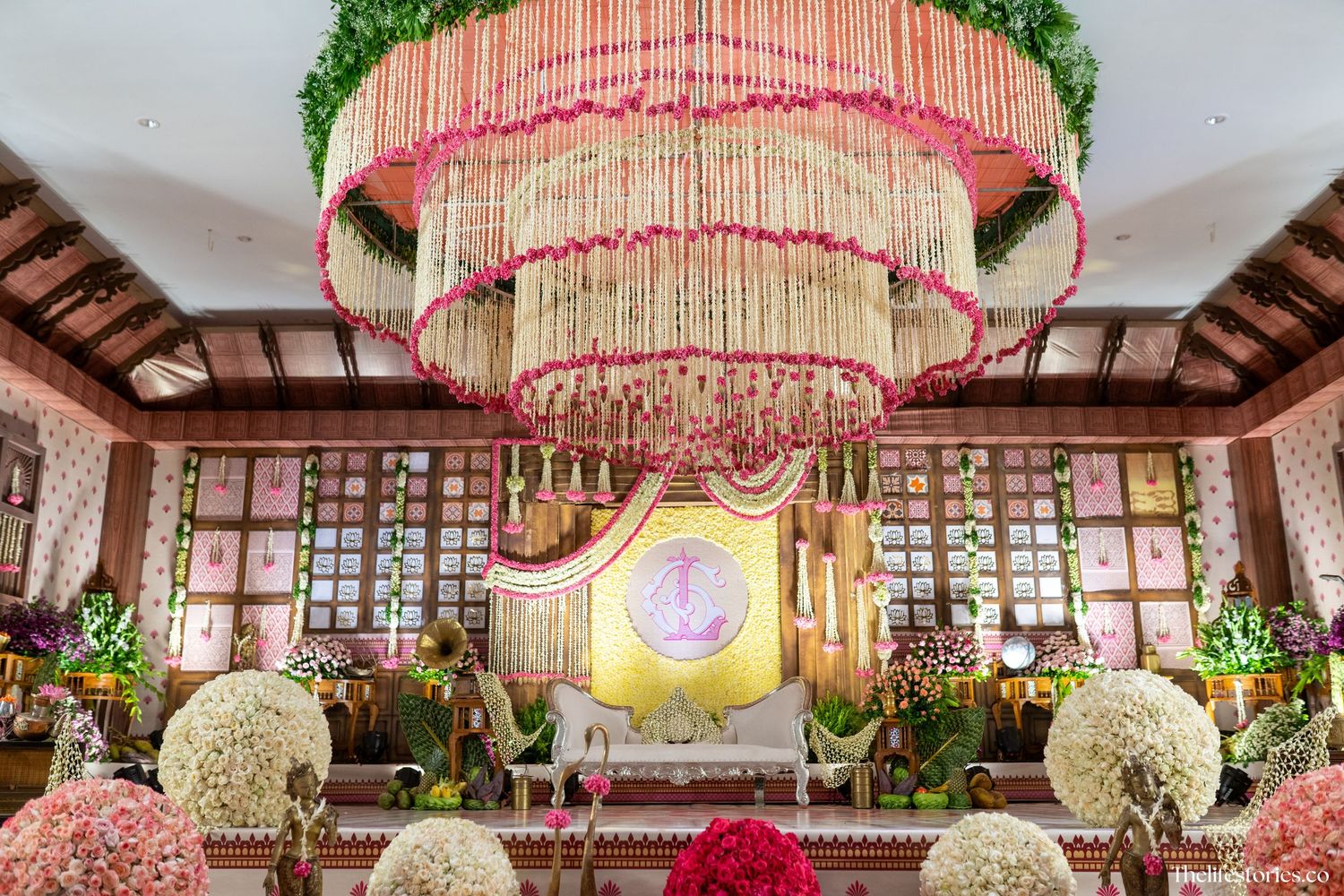Elegant wedding venue with three-tiered pink chandelier, floral arrangements, and ornate stage backdrop with golden accents.