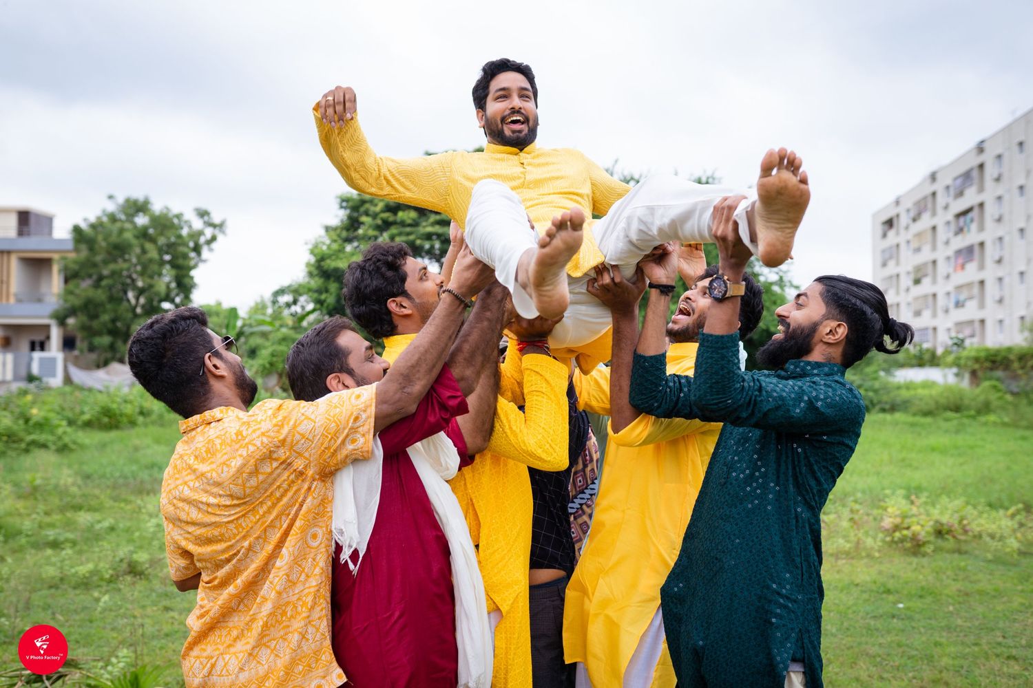 A group of friends in vibrant traditional Indian outfits lift and celebrate together in a grassy field.